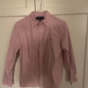 Rosa skjorta från Ralph Lauren - Snygg rosa skjorta från Ralph Lauren med klassisk krage och broderad logga på bröstet. Skjortan har långa ärmar och knappar framtill. Perfekt för en stilren look. Passar xs lite större