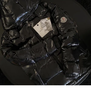 Svart pufferjacka från Moncler - Säljer en snygg svart pufferjacka från Moncler med glansig yta och huva. Jackan har dragkedja framtill och Moncler-logga på ärmen. Perfekt för kyliga dagar. Vet ej om den är äkta men nfc på loggan funkar och man kommer in på rätt hemsida!
