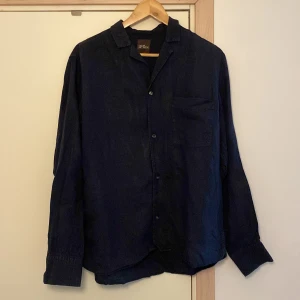Mörkblå skjorta från Oscar Jacobson - Snygg mörkblå skjorta från Oscar Jacobson i storlek M. Skjortan har långa ärmar och en klassisk krage med knappar framtill. Perfekt för en stilren look. Original pris 1500kr