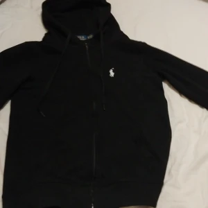 Polo-Ralph lauren kofta  - Säljer en klassisk svart hoodie från Polo Ralph Lauren. Den har en dragkedja framtill och en liten broderad logga på bröstet. Tillverkad i 100% bomull för en bekväm känsla. Perfekt för en stilren look.