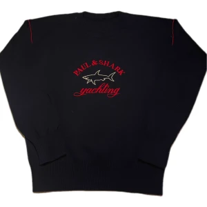 Svart tröja från Paul & Shark - Säljer en svart tröja från Paul & Shark med röd och vit broderad logga på framsidan. Tröjan har långa ärmar och är tillverkad i en blandning av ull och akryl. Perfekt för en stilren look.