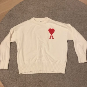 Vit tröja från Ami Alexandre Mattiussi - Säljer en stilren vit tröja från Ami Alexandre Mattiussi med ett rött hjärta och bokstaven A på bröstet. Tröjan har långa ärmar och ribbstickade muddar vid ärmslut och nederkant. Perfekt för en avslappnad och trendig look.