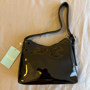 Svart axelväska från Silfen - Snygg svart axelväska i glansigt lack från Silfen. Väskan har en justerbar axelrem och stängs med dragkedja. Perfekt för att bära dina nödvändigheter med stil.