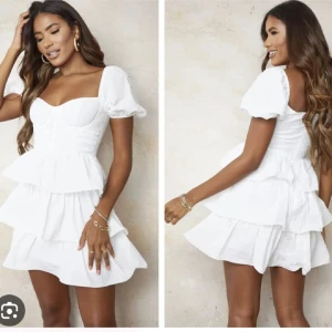 Vit kortklänning med volanger - Säljer en söt vit kortklänning med volanger och puffiga kortärmar. Klänningen har en smickrande passform med en v-ringad urringning och en omlott-design i midjan. Perfekt för sommarens alla tillfällen!