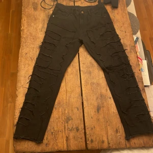 Svarta jeans med slitningar - Snygga svarta jeans med kraftiga slitningar längs benen. De har en klassisk femficksdesign och knappgylf. Perfekta för en all black look.