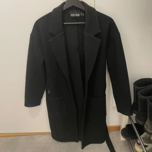 Svart kappa från Pieces - Snygg svart kappa från Pieces med klassisk design. Den har långa ärmar och ett bälte i midjan för en elegant passform. Perfekt för kyligare dagar. Hör av er om ni har frågor 🫶🏻
