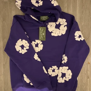 Lila hoodie med blommönster från Denim Tears - Säljer en cool lila hoodie från Denim Tears med ett unikt vitt blommönster. Hoodien har en klassisk passform med en stor ficka framtill och en bekväm huva. Perfekt för att sticka ut i mängden! Passar som M
