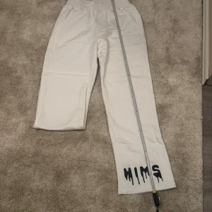 Vita mjukisbyxor med tryck - Snygga vita mjukisbyxor med elastisk midja och ett coolt svart tryck längst ner på ena benet. Perfekta för en avslappnad stil. 108 cm lång. Endast: 299