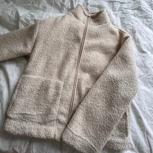 Beige teddyjacka - Mysig beige teddyjacka med dragkedja framtill. Jackan har en hög krage och två stora fickor. Perfekt för kyliga dagar när du vill hålla dig varm och stilren.
