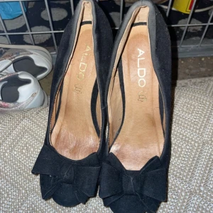 Svarta klackar från Aldo - Eleganta svarta klackar från Aldo med hög klack och dekorativ rosett framtill. Skorna har en klassisk design som passar perfekt för en stilren look.