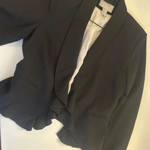 Kort kavaj med frill - Svart kortare kavaj med lite volang fram. Ger en mer dressad och feminin touch än vanlig rak linje. Väldigt snygg öppen över linne, tröja men lika bra med blus om man vill det såklart. Med kavajen knäppt får du det lilla extra med volangerna fram. 