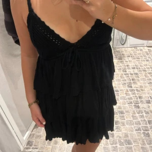 Svart klänning med volanger - Snygg svart klänning med volanger och knytning framtill. Den har en djup v-ringning och är ärmlös, vilket ger en elegant look. Perfekt för sommarens alla tillfällen.