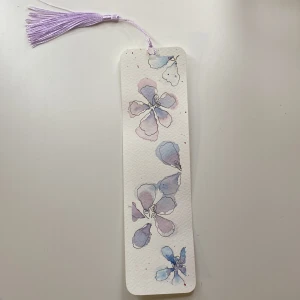 Bokmärke med blommotiv - Ett vackert handgjort bokmärke med akvarellmålade blommor i lila och blå nyanser. Bokmärket har en lila tofs upptill och är perfekt för att hålla koll på var du är i din bok. Tillverkat av tjockt papper för hållbarhet.
