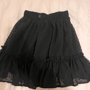 Svart kjol med volang - Snygg svart kjol med volangdetaljer och små prickar. Perfekt för att ge en chic touch till din outfit. Den har en elastisk midja för bekväm passform.