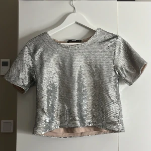 Silvrig paljettopp från BikBok - Säljer en glittrig silvrig topp från BikBok. Den är kortärmad och croppad, perfekt för en festlig kväll. Passar bra till både kjol och byxor.