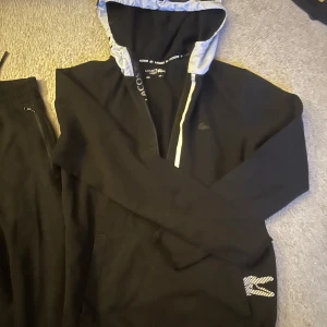 Svart tracksuit från Lacoste - Säljer ett par mjukis sätt  från Lacoste med vita detaljer. Perfekta för en avslappnad stil.