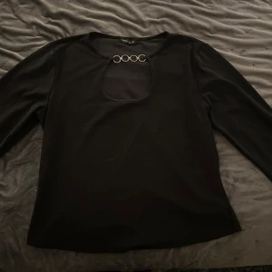 Svart topp från Vero Moda - Snygg svart topp från Vero Moda med unik detalj i form av fyra ringar vid halsen. Perfekt för en stilren look. Toppen har långa ärmar och en rund halsringning. Passar mig som vanligtvis bär S