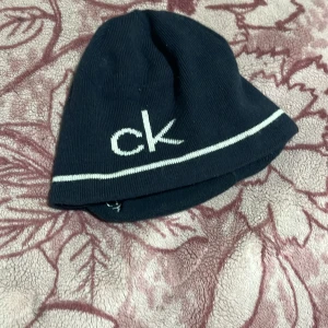 Svart mössa från Calvin Klein - Snygg svart mössa från Calvin Klein med vit broderad logga 'ck' framtill. Mössan har en enkel design med en vit rand som detalj nära kanten. Perfekt för att hålla värmen under kyliga dagar.