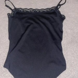 Svart linne med spetsdetaljer - Snyggt svart linne med smala axelband och spetsdetaljer längs halsringningen. Perfekt för lager-på-lager eller som det är. Enkelt och stilrent plagg för många tillfällen. Helt oanvända 