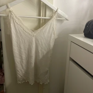 Vit linne med spetsdetaljer - Säljer ett stilrent vitt linne med v-ringad spetskant. Perfekt för varma dagar eller som ett lager under en tröja. Linnet har en enkel och elegant design.