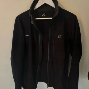 Svart jacka från Haglöfs - Säljer en svart jacka från Haglöfs med dragkedja framtill. Jackan har en stilren design och är perfekt för utomhusaktiviteter. Den har långa ärmar och en hög krage för extra skydd mot väder och vind.