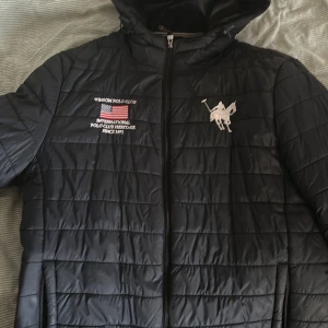 Svart quiltad jacka | Vinson Polo Club - Snygg svart quiltad jacka från Vinson Polo Club med dragkedja och huva. Jackan har broderade detaljer på bröstet med text och en flagga. Perfekt för kyligare dagar.