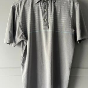 Grå pikétröja från FootJoy - Säljer en stilren grå pikétröja från FootJoy med diskreta vita ränder och tre knappar framtill. Perfekt för en avslappnad och sportig look. Tröjan har korta ärmar och en klassisk krage.