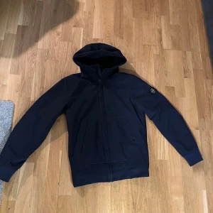 Blå windbreaker från Stone Island - Säljer en stilren blå windbreaker från Stone Island med dragkedja och huva. Den har långa ärmar och en klassisk passform. Perfekt för en avslappnad look.