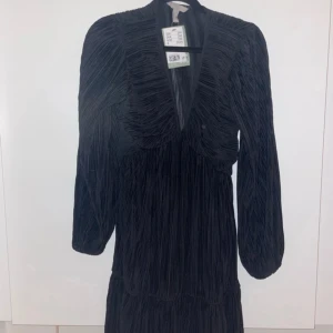 Svart klänning med v-ringning - Elegant svart klänning med v-ringning och långa ärmar. Klänningen har en fin struktur och är perfekt för en stilren look. Passar bra till både vardag och festliga tillfällen. Oanvänd med prislapp kvar. Org. Pris 299kr