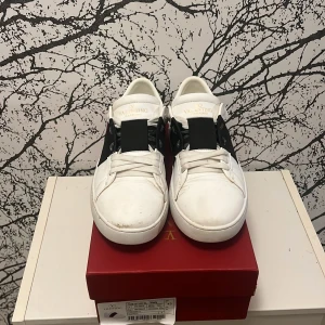 Valentino Opens  - Vita Valentino Opens med en bred svart rem över ovansidan. Skorna har en stilren design med snörning och en unik detalj på hälen. Snörena är lite slitna därav priset