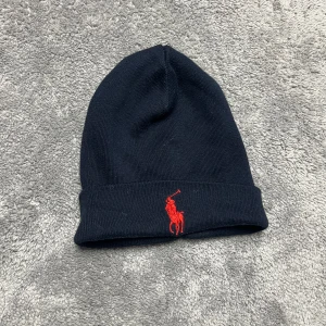 Mörkblå mössa från Ralph Lauren - Snygg mörkblå mössa från Ralph Lauren med röd broderad logga framtill. Perfekt för kyliga dagar och ger en stilren look. Tillverkad i mjukt material för extra komfort.