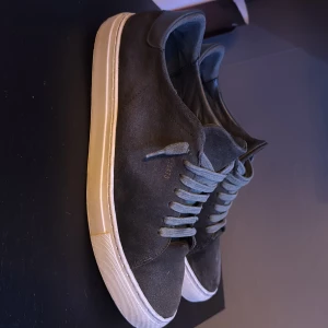 Grå sneakers från Axel Arigato - Snygga grå sneakers i mocka från Axel Arigato med vit sula och klassisk snörning. Perfekta för en stilren look.