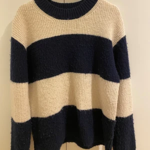 Randig stickad tröja från H&M - Mysig stickad tröja från H&M i beige och mörkblått med breda ränder. Perfekt för kyliga dagar med sin avslappnade passform och långa ärmar. En klassisk och stilren design som passar till många tillfällen.