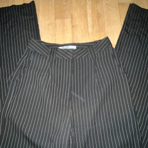 Svarta randiga kostymbyxor från nakd - Snygga svarta kostymbyxor från na-kd med vita ränder. Byxorna har en klassisk passform med dragkedja och knapp.