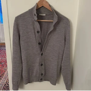 Grå kofta från Suitsupply - Snygg grå kofta från Suitsupply i merinoull. Den har en klassisk design med knappar framtill och ribbade muddar vid ärmslut och nederkant. Perfekt för lager-på-lager under kyligare dagar.