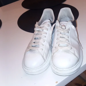 Vita sneakers från Alexander McQueen - Snygga vita sneakers från Alexander McQueen med svart detalj på hälen. Skorna har klassisk snörning och en chunky sula som ger en modern look. Perfekta för att lyfta vilken outfit som helst.