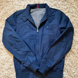 Blå jacka från J.Lindeberg - Snygg marinblå jacka från J.Lindeberg med dragkedja och ribbade muddar. Perfekt för en stilren look. Jackan har en liten broderad logga på bröstet och en klassisk krage. Storlek S i mycket bra skick 