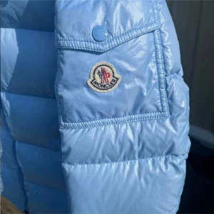 Ljusblå dunjacka från Moncler - Snygg ljusblå dunjacka från Moncler med dragkedja och knappar. Jackan har en hög krage och praktiska fickor med knäppning. Perfekt för kyliga dagar med stil.