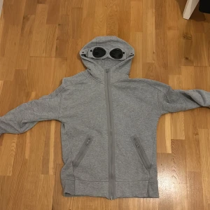 Grå huvtröja med glasögon från C.P. Company - Säljer en grå huvtröja från C.P. Company med inbyggda glasögon i huvan. Tröjan har dragkedja framtill och två fickor med dragkedjor. Perfekt för en unik och stilren look. Skick 8/10. Kvitto saknas men ingår original klistermärken. Storlek XS passar S. 