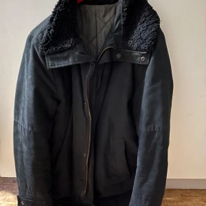 Svart jacka från Tiger of Sweden - Säljer en stilren svart jacka från Tiger of Sweden. Jackan har en mysig pälsfodrad krage och dragkedja framtill. Insidan är quiltad med märkets logotyp. Perfekt för kyligare dagar. Jackan är lång och går förbi midja