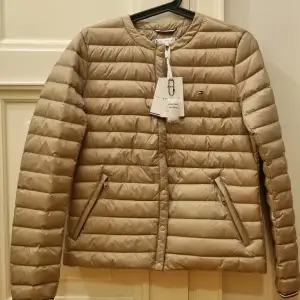 Helt ny Tommy hilfiger höstjacka i stl S.  Den är vattenavvisande och yttermaterial är polyester och fyllningen består av 90% dun och 10% fjäder. Lättviktsjacka som kan paketeras in i ett fack. Färg: medium taupe  