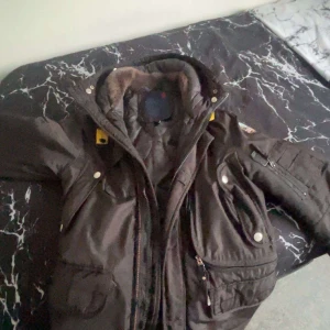 Brun vinterjacka med pälsfoder - Säljer en brun vinterjacka med pälsfoder. Den har en dragkedja och flera fickor med knappar. Jackan är perfekt för kalla vinterdagar och har en cool patch på ärmen. Den är i bra skick och redo för nya äventyr!