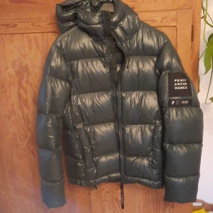 Grön pufferjacka från Peak Performance - Säljer en snygg och varm grön pufferjacka från Peak Performance. Jackan har en glansig yta och är perfekt för kalla vinterdagar. Den har en dragkedja framtill och en praktisk huva. På ärmen finns en cool logga . Jackan är lagad på ett ställe. Se bild