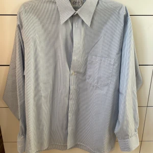 Randig skjorta från Oscar of Sweden - Säljer en stilren och klassisk skjorta från Oscar of Sweden. Skjortan är i strykfri bomull, vilket gör den superlätt att sköta. Den har en snygg blå och vit randig design med långa ärmar och en bröstficka. Perfekt för både kontoret och mer avsl