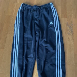 Svarta träningsbyxor från Adidas - Säljer ett par svarta träningsbyxor från Adidas med de klassiska tre ränderna längs sidorna. Byxorna har en elastisk midja med snörning för bästa passform. Perfekta för träning eller en avslappnad dag hemma. De är i bra skick och redo för nya äventyr!