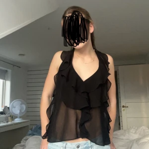 Svart ärmlös blus med volanger - Snygg svart blus i transparent material med volanger framtill. Perfekt för en utekväll eller fest! Den är ärmlös och har en v-ringad design som ger en elegant touch. Passar bra till både jeans och kjol.