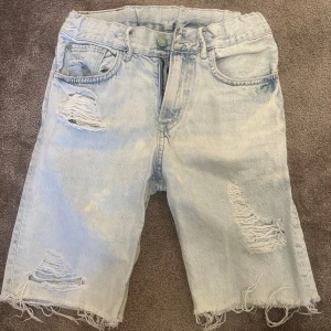 Ljusa jeansshorts med slitningar - Säljer ett par ljusa jeansshorts i slim fit med coola slitningar. Perfekta för sommaren! De har en klassisk femficksdesign och är gjorda i denim. Shortsen är i bra skick och redo för nya äventyr. Passar perfekt till en avslappnad stil.
