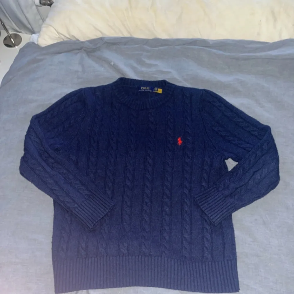 Säljer en snygg mörkblå stickad tröja från Ralph Lauren med det klassiska röda logotypbroderiet på bröstet. Tröjan har ett kabelstickat mönster och långa ärmar, perfekt för kyligare dagar. Passar både till jeans och chinos!. Neuletakit & Villapaidat.