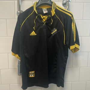Svart och gul fotbollströja från Adidas - Retro aik tröja. Snygg svart fotbollströja från  Tröjan har korta ärmar och en klassisk krage med snörning. Perfekt för fotbollsfans som vill visa sitt stöd. Försöker frakta samma dag 