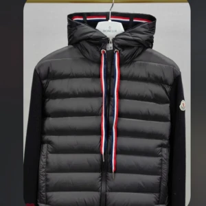 Moncler cardigan  - Snygg svart pufferjacka från Moncler med långärmade stickade ärmar. Jackan har en dragkedja i rött, vitt och blått samt en hög krage. Perfekt för kyligare dagar.